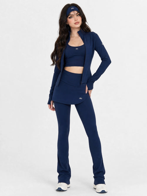 A woman modeling a matching navy blue activewear set consisting of a cropped tank top, a short jacket, and high-waisted flared pants, paired with white sneakers against a light gray studio background.