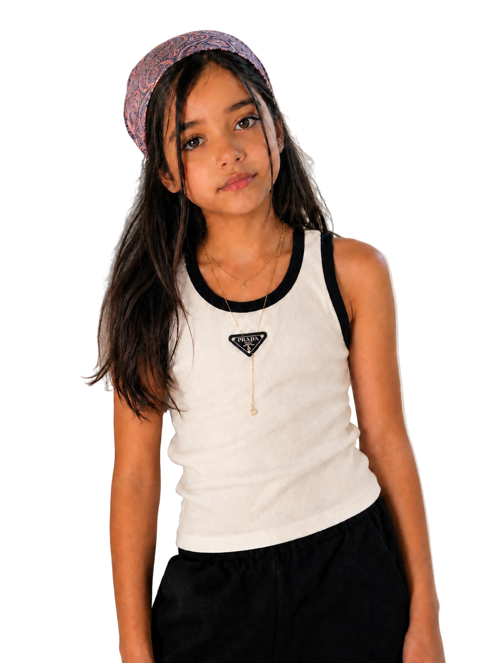 A young girl with long dark hair wearing a patterned headband, a white ribbed tank top with black trim, and black pants, leaning against a textured light beige wall.