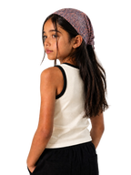 A young girl with long dark hair wearing a patterned mauve and blue paisley headband, a white ribbed tank top with black trim, and black pants, photographed against a neutral light gray textured background.