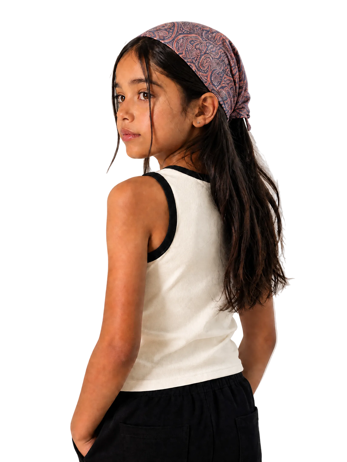 A young girl with long dark hair wearing a patterned mauve and blue paisley headband, a white ribbed tank top with black trim, and black pants, photographed against a neutral light gray textured background.