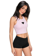 A woman modeling a light pink ribbed crop top with a black triangular logo patch on the chest, paired with black athletic shorts, against a neutral gray background.