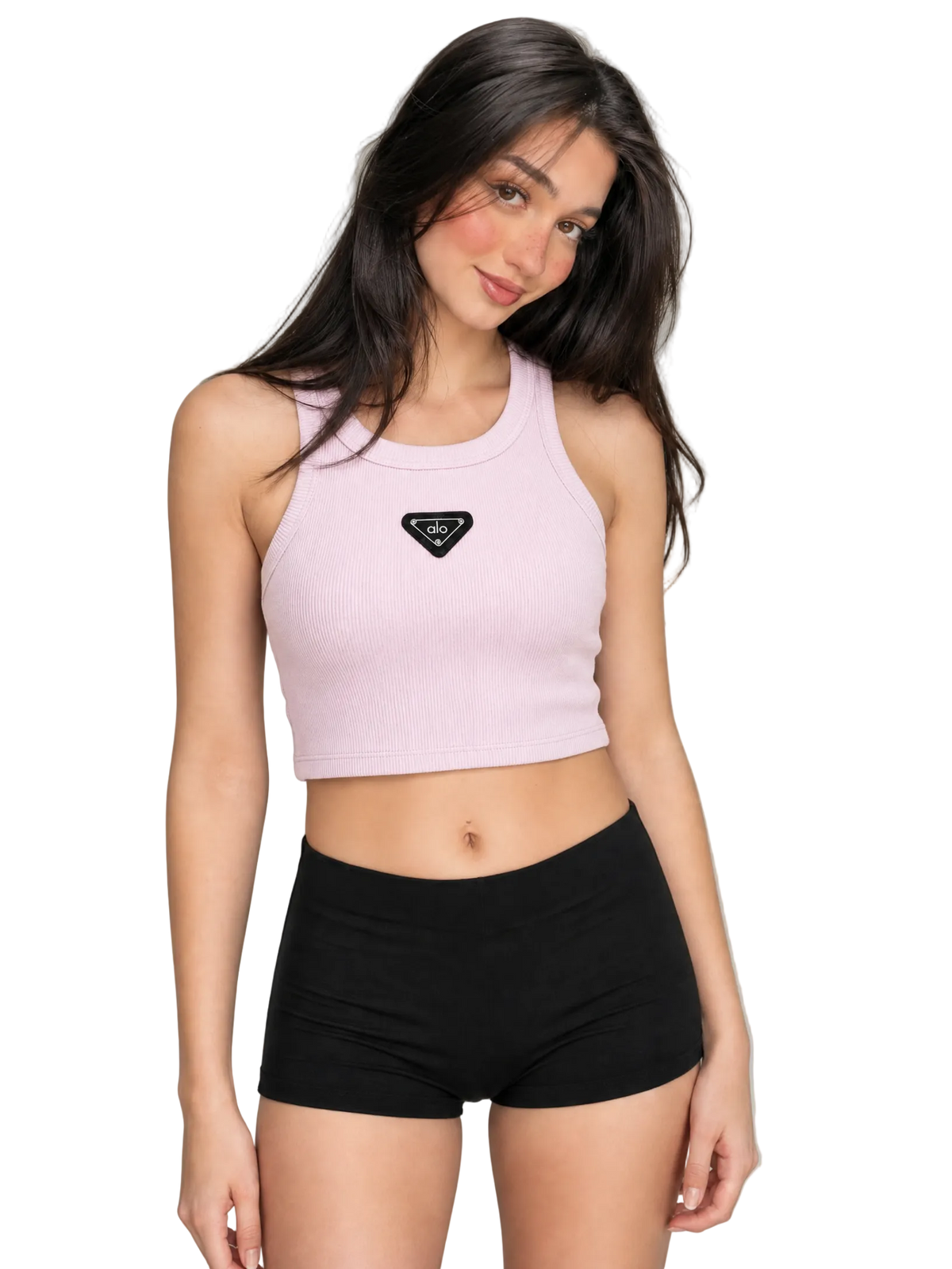 A woman modeling a light pink ribbed crop tank top with a black triangular logo patch on the chest, paired with black athletic shorts, against a neutral gray studio background.
