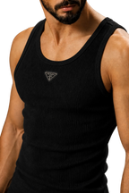 A close-up of a man wearing a black ribbed tank top with a small triangular logo patch on the chest, photographed against a neutral gray studio background.