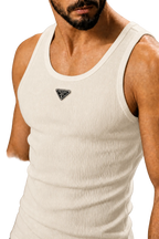 A close-up of a man wearing a ribbed cream-colored tank top with a small black triangular logo patch on the chest, set against a neutral gray background.