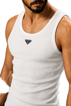 A close-up of a man wearing a white ribbed tank top with a small black triangular logo patch on the chest, set against a neutral gray background.