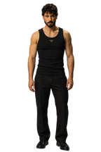 A man modeling a black sleeveless tank top with a small silver logo on the chest, paired with black trousers and black shoes, against a neutral gray studio background.