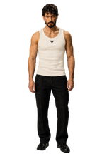 A man modeling a white ribbed tank top with a small black Prada triangle logo on the chest, paired with black trousers and black shoes, standing against a textured gray studio background.