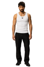 A man modeling a white ribbed tank top with a small black Prada triangle logo on the chest, paired with black trousers and black shoes, against a neutral gray studio background.