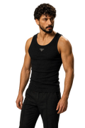 A muscular man with curly dark hair and a beard wearing a black ribbed tank top with a small silver geometric logo on the chest, paired with black pants, against a neutral gray studio background.
