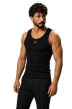 A muscular man with curly dark hair and a beard wearing a black ribbed tank top with a small silver geometric logo on the chest, paired with black pants, against a neutral gray studio background.