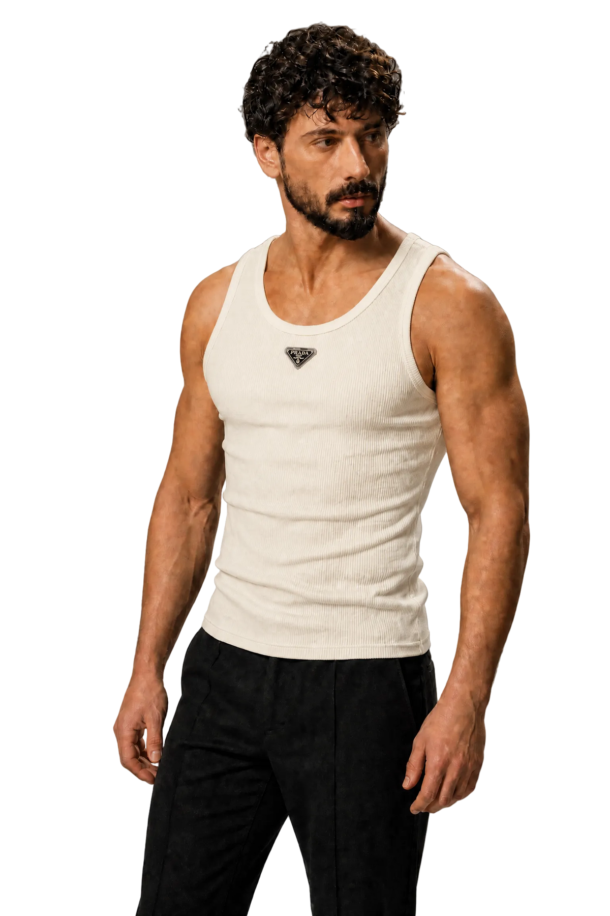 A muscular man with curly dark hair and a beard wearing a fitted off-white ribbed tank top with a small black geometric logo on the chest, paired with black pants, standing against a textured gray studio background.