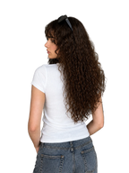 A woman with long, curly brown hair is shown from behind wearing a fitted white short-sleeve t-shirt and blue denim jeans in a modern living room setting.