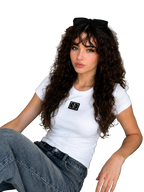 A woman with long, curly brown hair sits on a light gray couch, wearing a fitted white short-sleeve t-shirt with a black rectangular patch on the chest, blue jeans, and black sunglasses resting on her head.