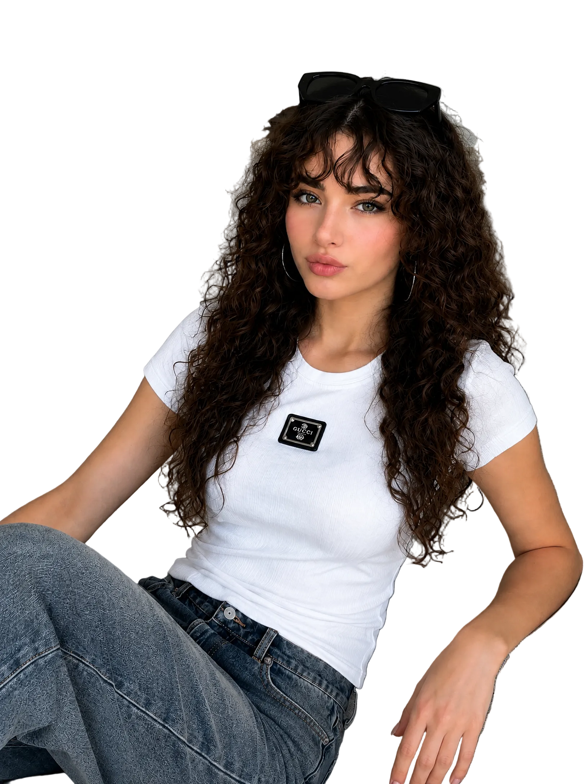 A woman with long, curly brown hair sits on a light gray couch, wearing a fitted white short-sleeve t-shirt with a black rectangular patch on the chest, blue jeans, and black sunglasses resting on her head.