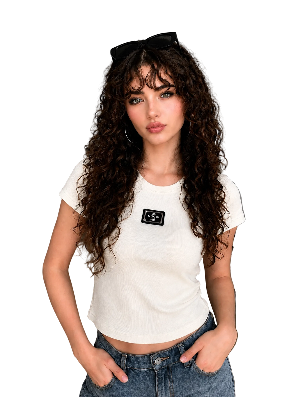 A woman with long, curly brown hair wearing a fitted white short-sleeve crop top with a small black chest patch and blue jeans, standing in a modern living room setting.