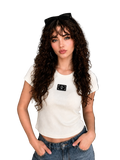 A woman with long, curly brown hair wearing a fitted white short-sleeve crop top with a small black chest patch and blue jeans, standing in a modern living room setting.