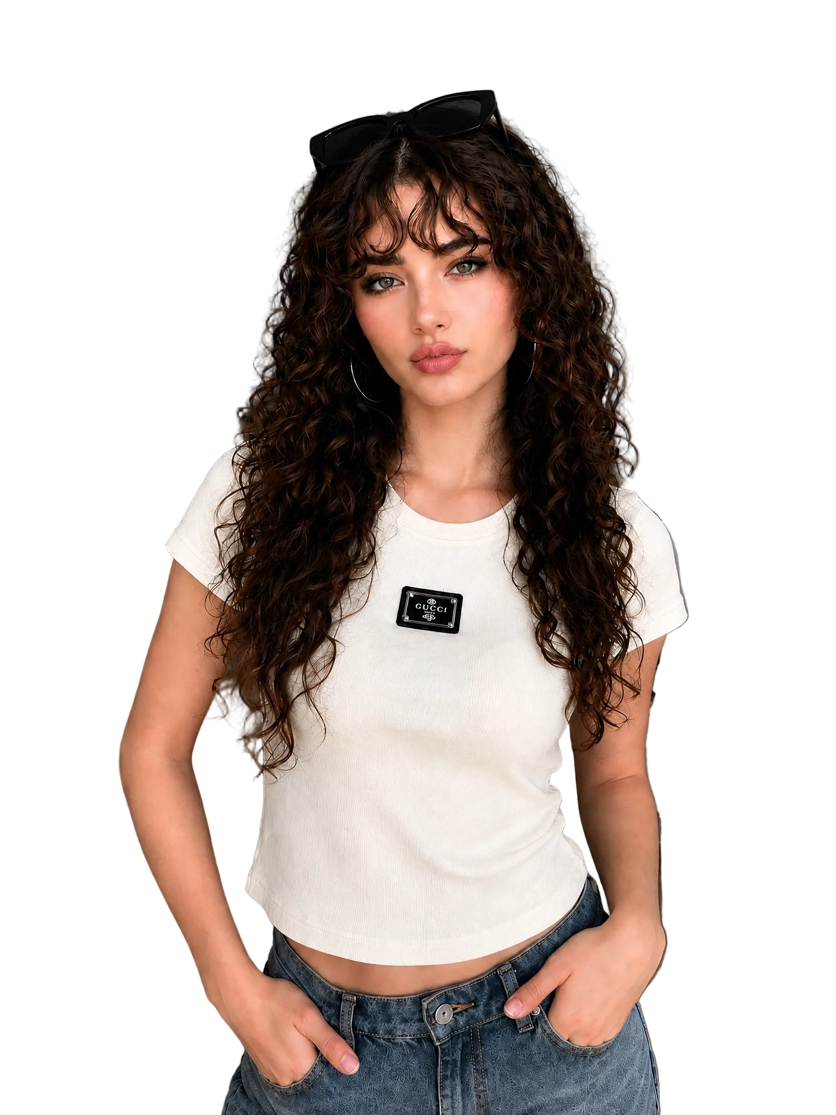 A woman with long, curly brown hair wearing a fitted white short-sleeve crop top with a small black chest patch and blue jeans, standing in a modern living room setting.