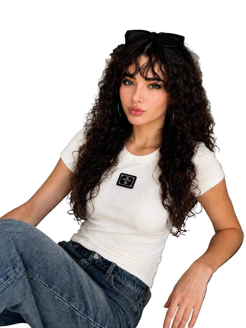 A woman with long, curly dark hair sits on a gray sofa wearing a fitted white short-sleeve t-shirt with a black rectangular patch on the chest, paired with blue jeans. She has black sunglasses resting on her head.
