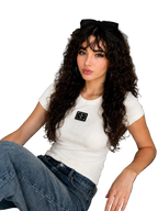 A woman with long, curly dark hair sits on a gray sofa wearing a fitted white short-sleeve t-shirt with a black rectangular patch on the chest, paired with blue jeans. She has black sunglasses resting on her head.