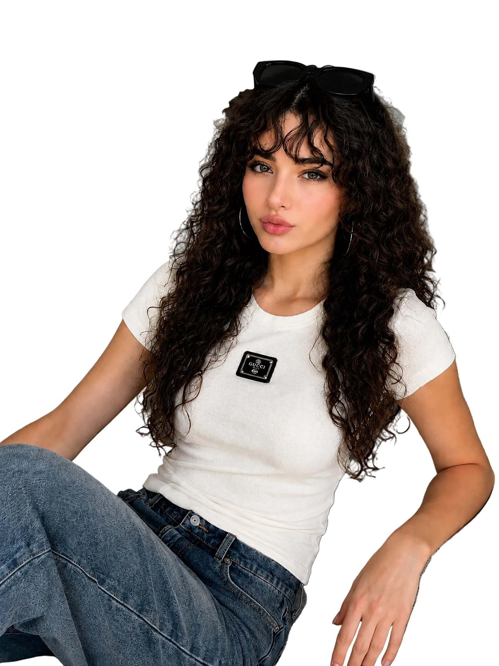 A woman with long, curly dark hair sits on a gray sofa wearing a fitted white short-sleeve t-shirt with a black rectangular patch on the chest, paired with blue jeans. She has black sunglasses resting on her head.