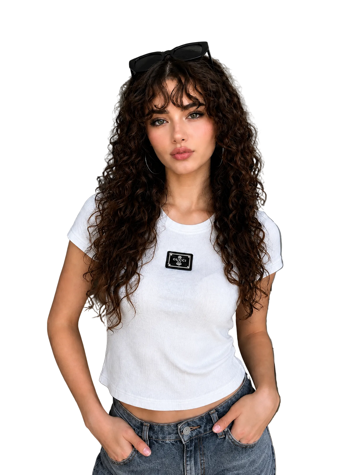 A woman with long, curly brown hair wearing a white short-sleeve crop top with a small black rectangular patch on the chest, paired with blue jeans. She is standing in a modern living room setting.