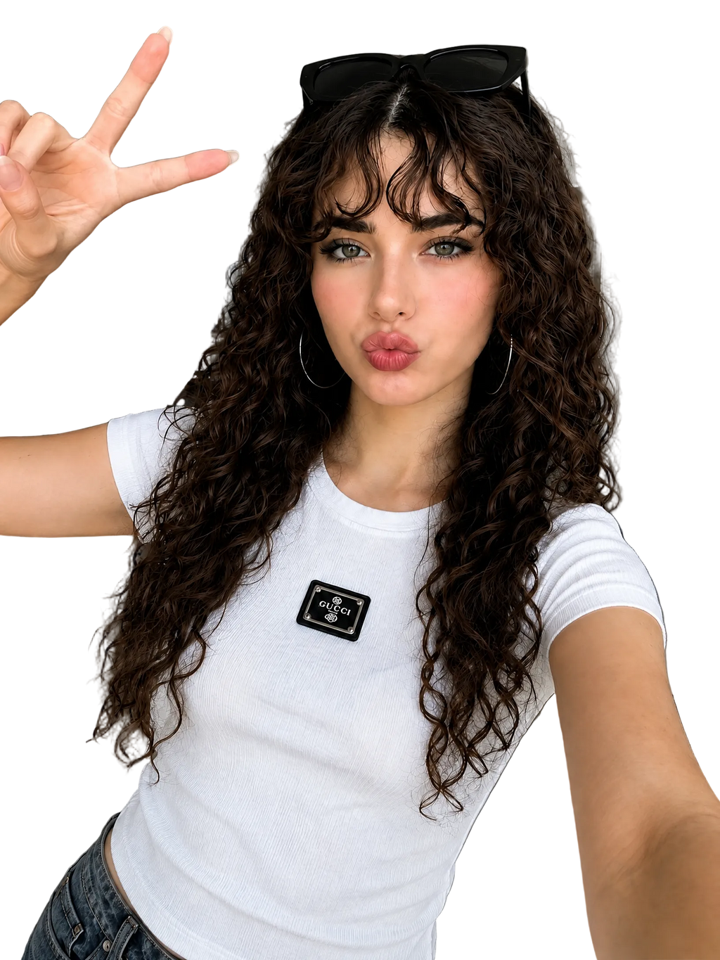 A woman with long, curly brown hair takes a selfie in a modern apartment. She is wearing a white short-sleeve t-shirt with a black rectangular patch on the chest, blue jeans, and black sunglasses resting on her head.