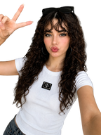 A woman with long, curly brown hair takes a selfie in a modern apartment. She is wearing a white short-sleeve t-shirt with a black rectangular patch on the chest, blue jeans, and black sunglasses resting on her head.