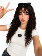 A woman with long, curly brown hair wearing a white ribbed short-sleeve t-shirt with a black rectangular patch on the chest, posing in a modern living room setting.