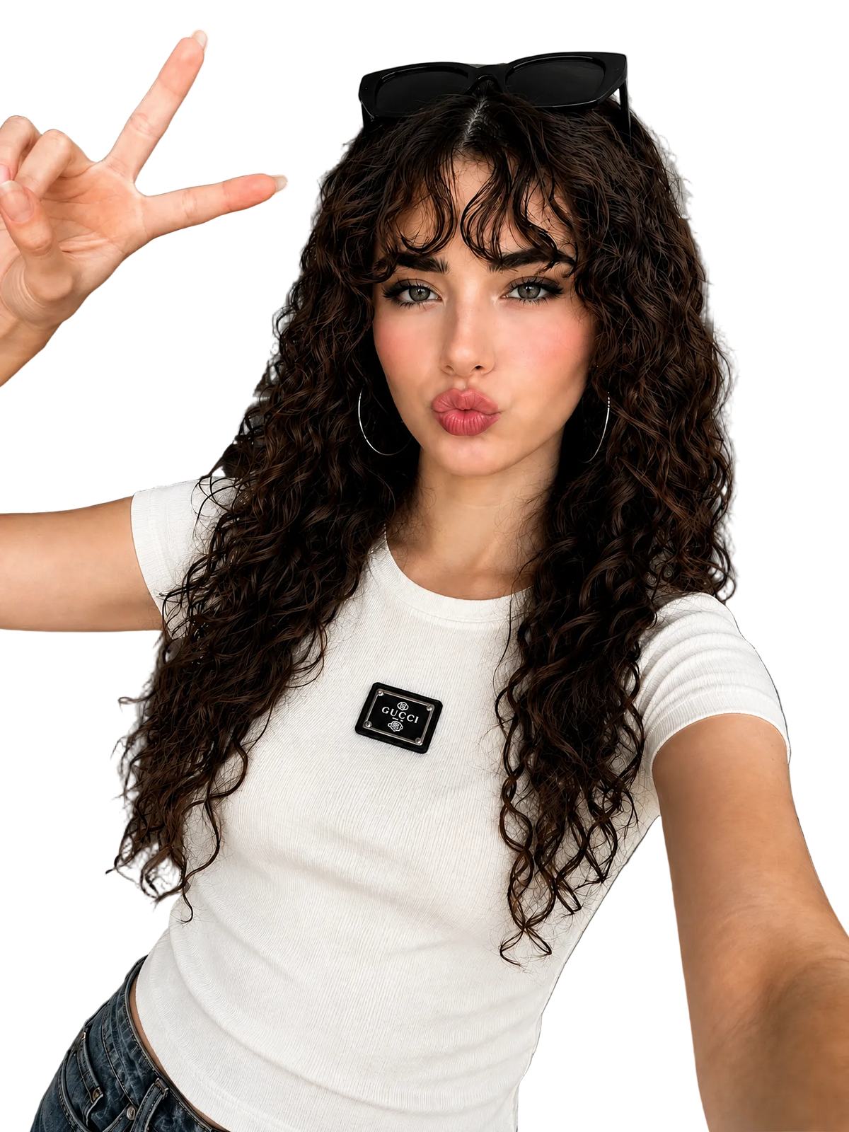 A woman with long, curly brown hair wearing a white ribbed short-sleeve t-shirt with a black rectangular patch on the chest, posing in a modern living room setting.